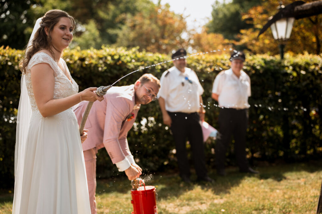 Im Vordergrund löscht Brautpaar ein Feuer. Im Hintergrund sind Feuerwehrmänner