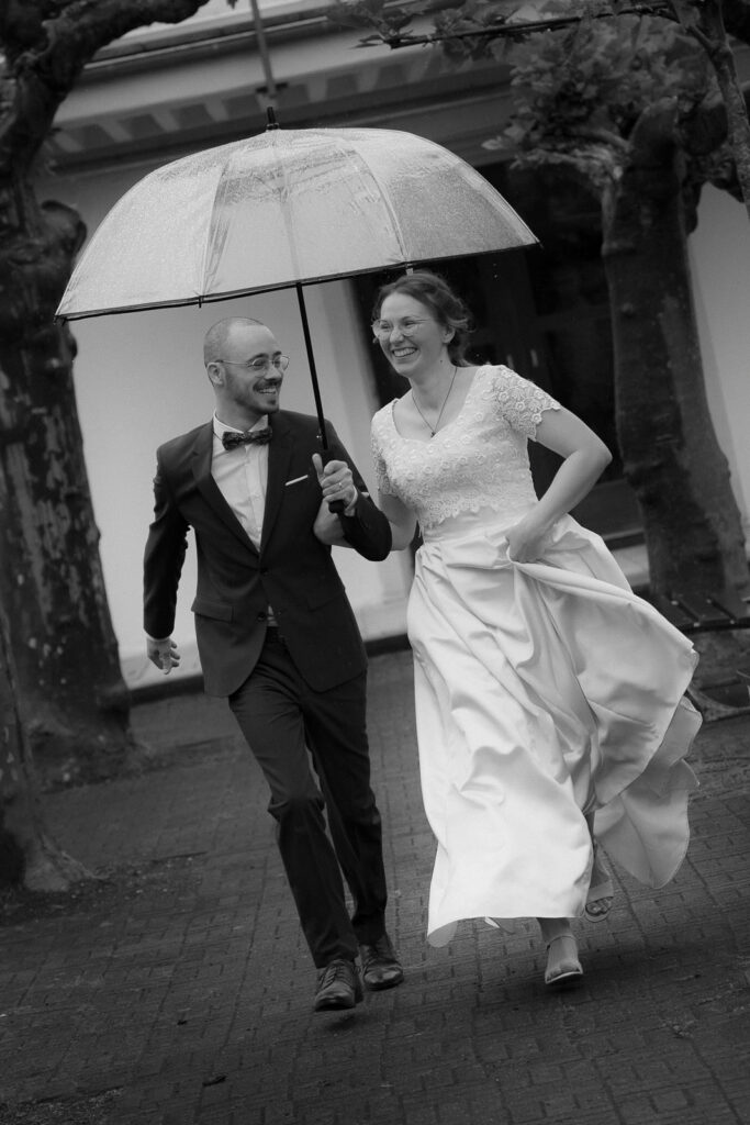 Brautpaar läuft mit Regenschirm auf Fotografin zu