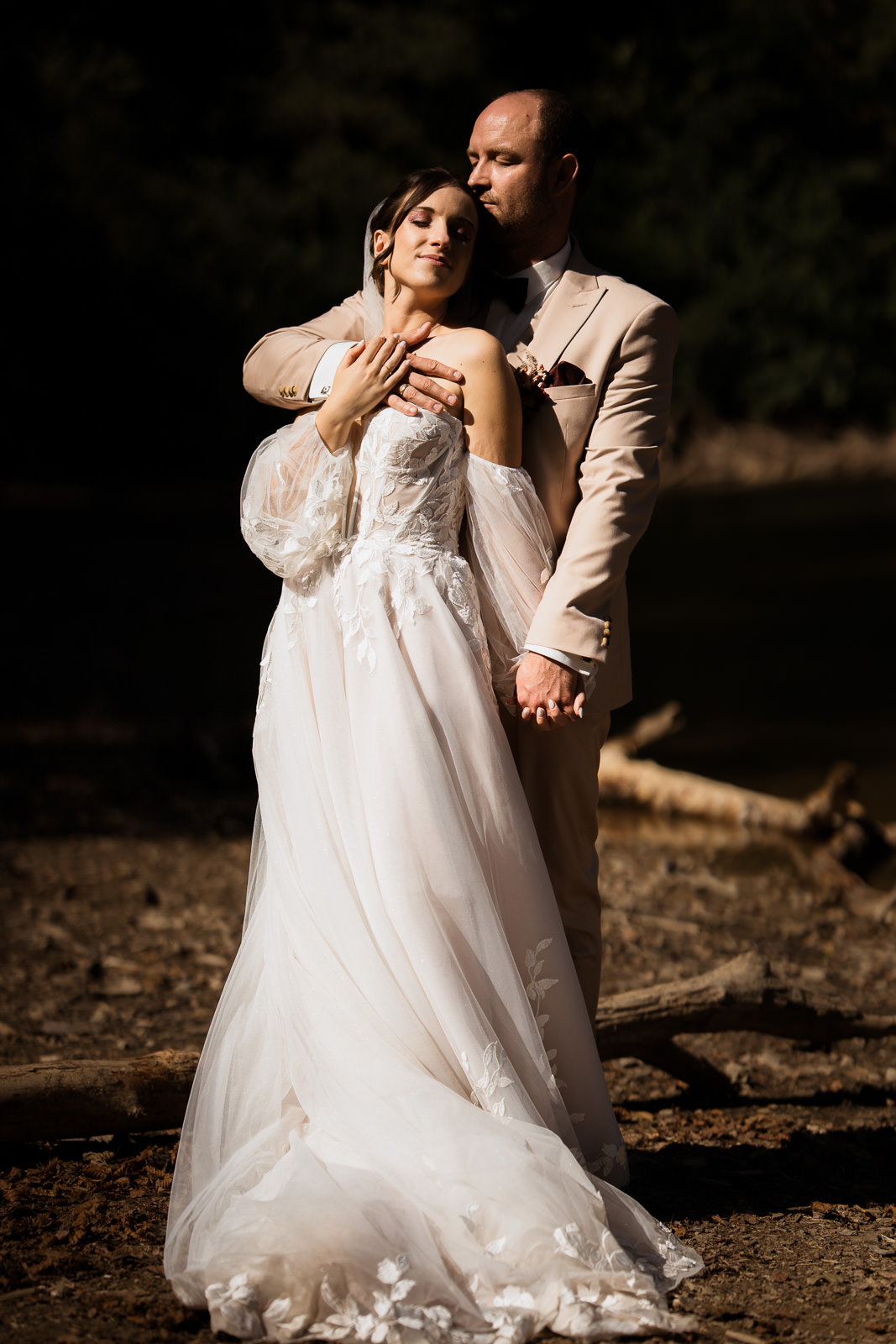 Braut steht mit dem Rücken an den Bräutigam gelehnt, beide lehnen sich leicht zurück im Sonnenlicht, dunkler Hintergrund