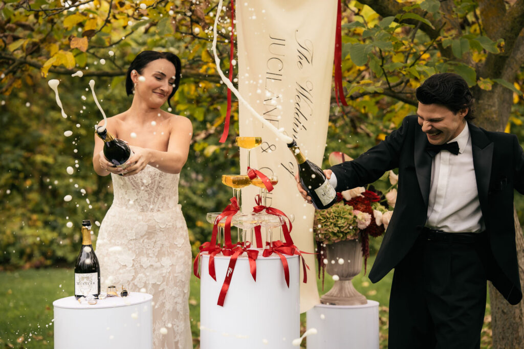 Brautpaar mit Champagnerturm hat gerade zwei Champagnerflaschen geöffnet. Der Champagner spritzt.
