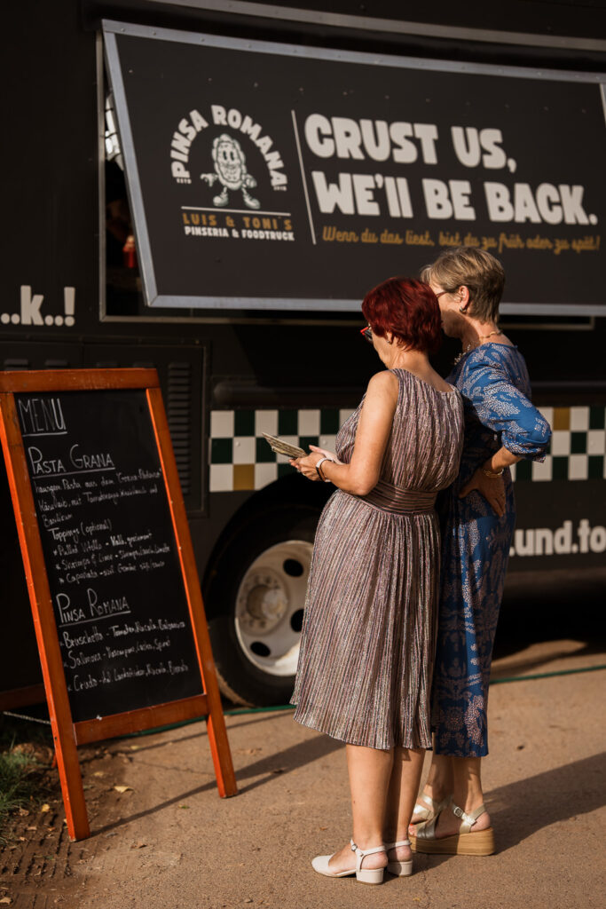 Food Truck mit geschlossener Klappe, zwei Frauen stehen davor und lesen die Speisekarte auf einer Tafel