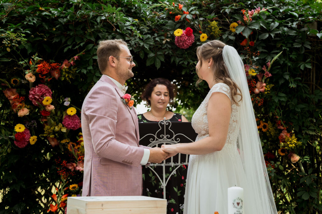 Brautpaar steht sich bei einer freien Trauung gegenüber, im Hintergrund ist die Traurednerin zu sehen