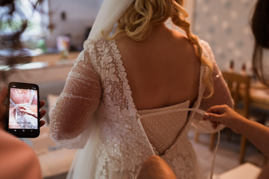 Trauzeugin schnürt das Brautkleid an der Braut zu. Fotografin zeigt das im Rahmen der getting ready Reportage