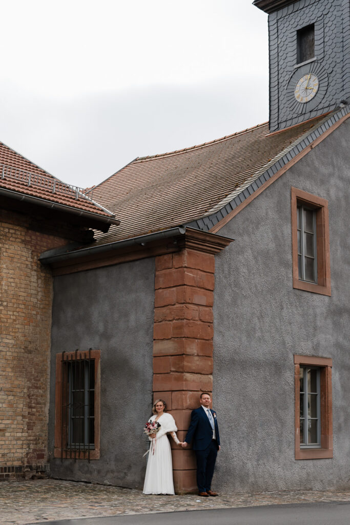 Brautpaar steht genenseitig Hände haltend um die Ecke eines Hauses.