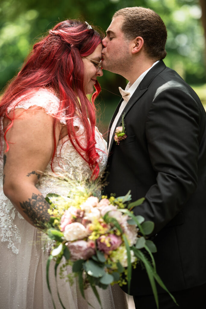 Rothaarige Braut wird von Bräutigam auf Stirn geküsst