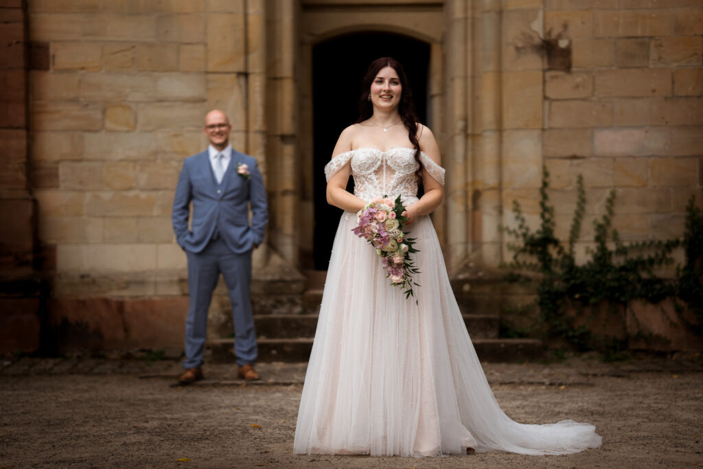 Braut schaut Fotografin an, Bräutigam ist unscharf hinten im Hintergrund zu sehen schaut ebenfalls Richtung Kamera
