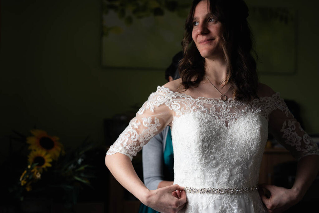 Braut schaut zum Fenster während ihre Mutter ihr Kleid schließt