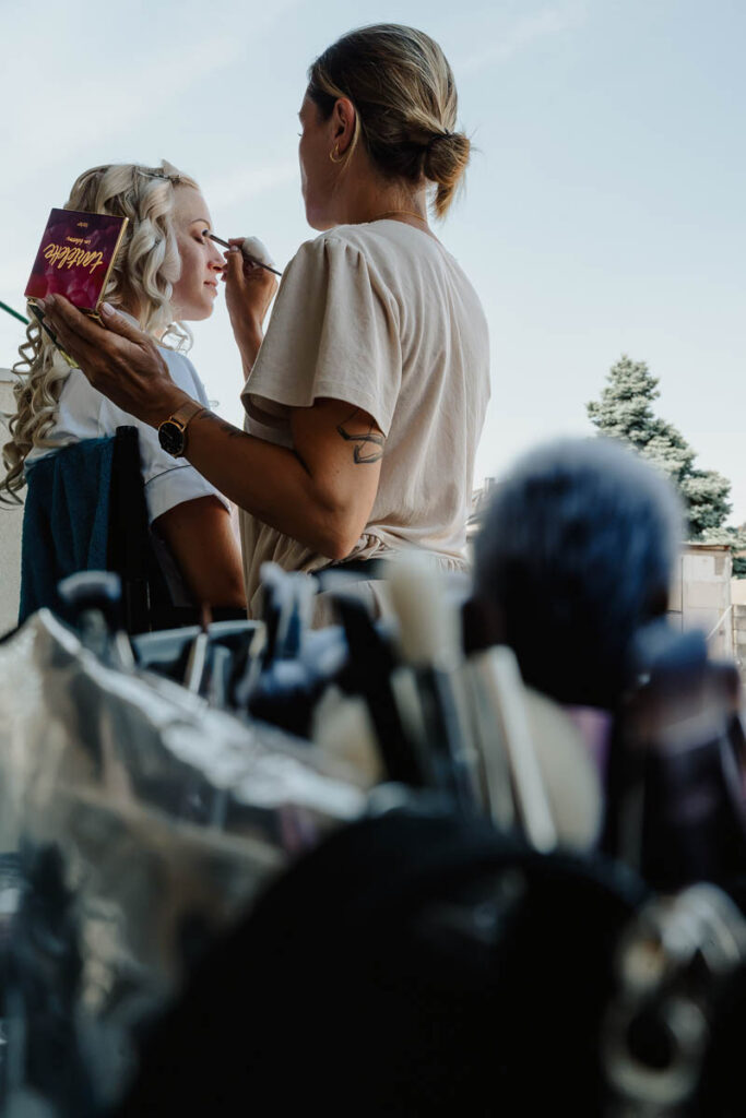 Braut wird von einer Brautstylistin geschminkt. Fotograf wählt Perspektive mit Schminkutensilien im Vordergrund