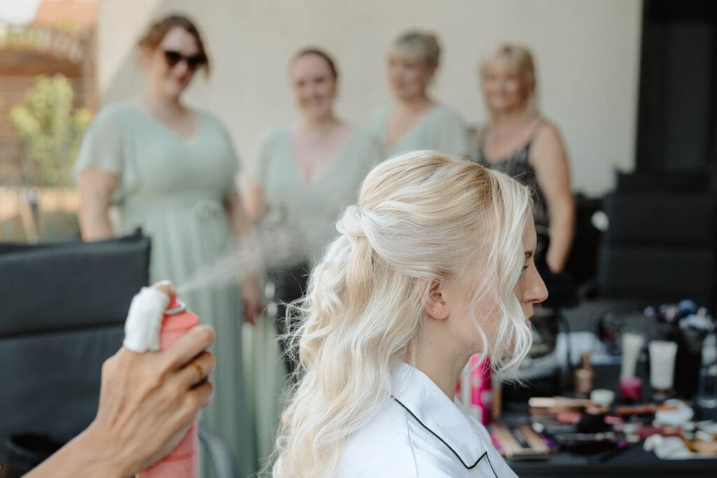Im Hintergrund Brautjungfern im Vordergrund Braut und die Stylistin spüht Haarspray